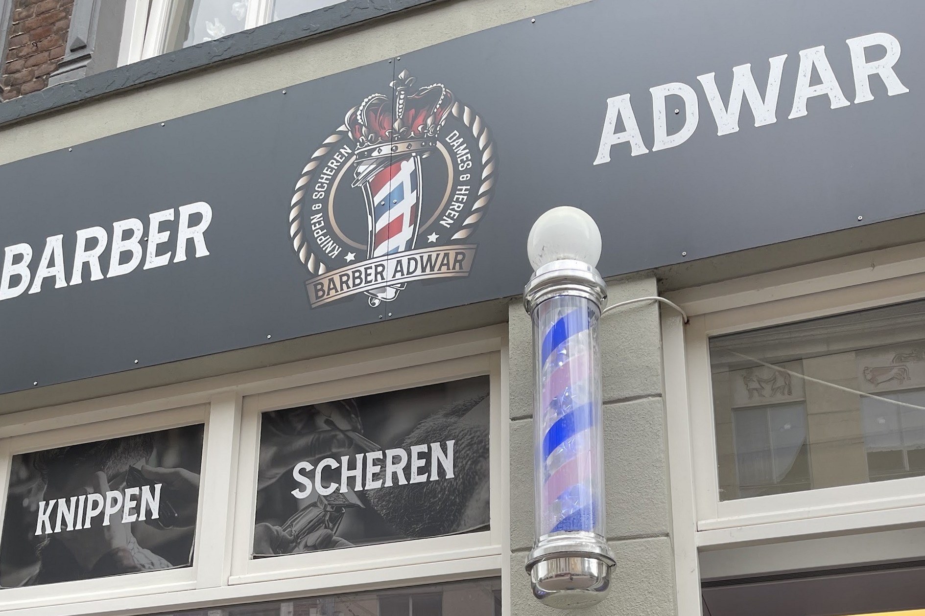 De winkelpui van Barber Adwar aan de Hamstraat, met het logo op de gevel, Knippen en Scheren op de ramen en de kapperspaal naast de deur